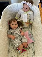 Photo showing two reborn dolls lying side by side in a white basket with quilted lining, one dressed in pajamas with a headband and the other in a polka dot outfit and socks with a pink headband.