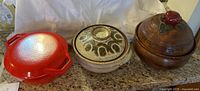 Three covered casserole dishes: red enameled pot, beige ceramic pot with brown pattern, brown ceramic pot with apple-shaped lid knob.