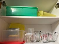 Four glass measuring cups and several plastic storage containers in a shelving unit. Colors include green, yellow, clear, and red lids.