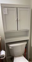 Photo of a white wooden bathroom shelf sitting over a toilet, featuring two doors on the upper cabinet and an open shelf below.
