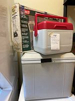 Electric portable fridge on bottom with small red and white cooler on top. Also a boxed Coleman propane stove behind the cooler.