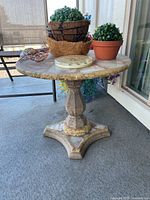 Full view of the cast marble side table on a patio with plants and a decorative clock on top, showing the round marble top and pedestal base.