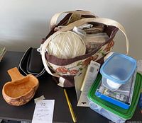 View of floral fabric bag containing yarn and sewing accessories, wooden scoop, and some small black trays.