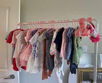 Wide view of all doll clothes hanging on pink hangers on a white clothing rack against a white wall and door