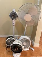 Five assorted cooling fans of various sizes and designs gathered on wooden floor against beige wall.