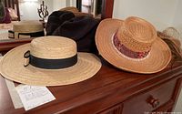 Photo showing multiple straw hats with different brims and bands, plus black fabric hats on a wooden dresser.