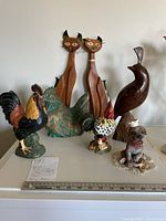 Full view of all figurines displayed on a surface showing two tall carved wooden cats, three roosters (two painted and one wooden), a solid polished wood bird, and a sand cast dog figurine with chef hat.