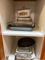 Upper cabinet shelf with three CorningWare casserole dishes with lids (floral and blue pattern) stacked and multiple glass baking pans including clear rectangular pans and a metal loaf pan.