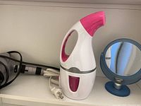 Pink and white handheld garment steamer and black curling iron with white power cord and small blue round mirror on shelf