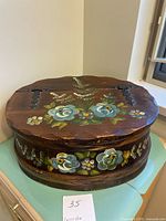 Side view of the oval wooden box showing floral hand painted decorations and hinges.