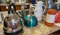 Lot overview showing electric kettle, stove top kettle, white ceramic thermos, and plastic thermos on countertop