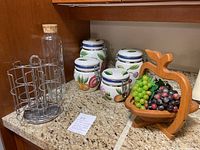 Wide view showing four fruit-pattern ceramic jars, wooden pear-shaped fruit stand with artificial grapes, metal mug holder carousel, and tall glass jar with cork stopper on kitchen counter.