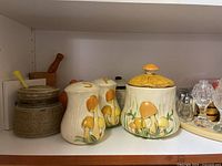 Shelf containing mushroom pottery items, including honey pot and numbered containers, with a small stoneware pot with a wooden dipper on left side.