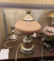 Photo displaying table lamp with silver metal base and pinkish ceramic center with no shade on, surrounded by several other lamps including frosted glass shade lamps and a floral painted shade lamp.