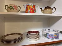 Shelf display showing Wade Fine China burgundy and gold patterned dessert bowls and salad plates, vintage orange flower pitcher, brown and beige teapot, dog-themed bowl with bone motifs