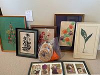 Six framed botanical artworks including embroidery and prints arranged on carpet, with embroidery hoop in plastic bag