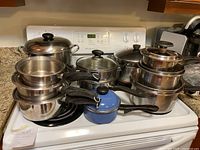 Stacked pots including small, medium, and large stainless steel pots with glass lids on kitchen stove. Small blue pot with lid in front.