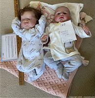 Two reborn dolls lying on a pink blanket, one with brown hair and asleep expression, the other bald with eyes open and wearing clothes