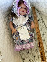 Full view of reborn doll lying down, showing floral dress, purple bonnet, and fixed limbs. Ruler next to doll for size reference.