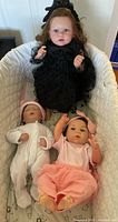 Photo showing three reborn dolls arranged in a lined bassinet or cradle; one large doll in black dress, two smaller dolls in pink and white baby clothing.