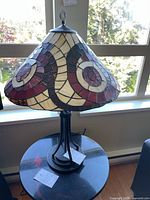 Full view of stained glass lamp with shade showing colorful geometric circular pattern and solid metal base on black round table by window.