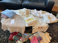 Overview of entire lot spread across two tables showing various linens including crochet lace tablecloths, embroidered cloths, gingham fabrics, and seasonal runners and towels.