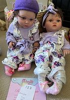 Photo shows pair of twin reborn dolls seated on carpet, one wearing purple knit hat, the other lavender headband. Both in floral overalls and lavender tops.