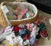 Wicker bassinet filled with assorted doll accessories including clothes and bottles, with more doll clothes spread on the carpet below.