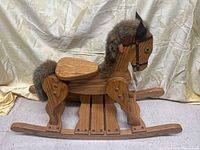 Side view of the wooden rocking horse showing the full body, seat, rocker rails, legs, faux fur mane and tail, green eyes, and bridle.