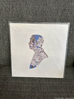 Front cover of white vinyl LP with silhouette cutout showing floral artwork inside, placed on gray fabric background.