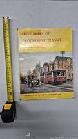 Front cover of the vintage TTC history book with a yellow background and illustration of streetcars and old cars, next to a tape measure showing approx 11 inch height