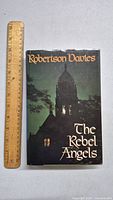 Front cover of The Rebel Angels book with ruler for size comparison showing mostly black and dark green colors with gothic tower image.