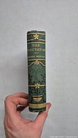 Spine of the antique book The Spectator showing title, author name Henry Morley, and decorative gold floral and geometric details on green cloth.