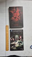 Two hardcover DC Comics comic books, one titled Injustice: Gods Among Us and the other with dark cover art, placed side by side with a ruler for size reference.