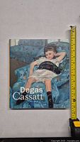 Front cover of art book showing a young girl reclining in a blue armchair, titled 'Degas Cassatt'. A tape measure alongside shows height approximately 10 inches.