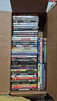 Top view of a box filled with 38 assorted DVD movies stacked vertically and horizontally, titles visible on covers and spines