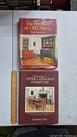 Photo showing both books stacked on a white surface with a ruler beside them for size reference. The top book is 'The Furniture of Old Ontario' by Philip Shackleton with a red dust jacket featuring a traditional furniture room image.