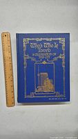 Front cover of the book showing blue hardcover with gold embossed title and city illustration next to a wooden ruler to indicate size.