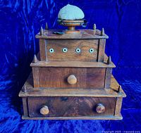 Front view of the wooden 3-tiered sewing box showing three drawers with wooden knob handles, bovine material eyelets, and pointed wooden finials at corners. Blue pincushion atop the rotating lid with visible wear on the black silk base.
