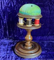 Front view of antique English sewing stand showing green silk pincushion on top and spindles with thread below on turned mahogany wood base.