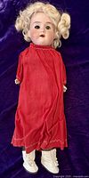 Full view of the doll dressed in red linen frock with blonde curly wig and white shoes, sitting on purple background.