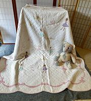 Full view of white quilted coverlet with ballerina embroidery and scalloped pink edging draped over screen with patchwork teddy bear seated on carpet in front.