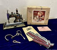 Full lot showing Casige Child's Sewing Machine 1015, original box, paperwork/manual, screwdriver, bodkin, clamp, and fabric pouch on dark blue surface.