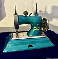 Front and side view of the teal Casige miniature child's sewing machine showing the hand crank and eagle logo.