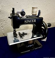 Front and side view of the black cast metal Child’s Singer SewHandy #20-10 sewing machine with spool of thread and additional accessories on the table.