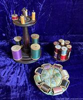 Photo showing four distinct thread holders arranged on a blue velvet surface. Walnut wooden thread stand holding several spools of thread, resin poker chip style thread caddy with six wells and multiple thread spools, handmade floral fabric pincushion with thread pockets.