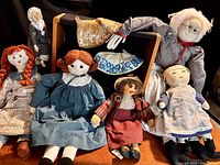 Five soft dolls in aprons and petticoats arranged sitting and standing showing yarn braids, hand-stitched features, and wooden painted head doll alongside two beaded bonnets.