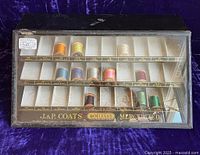 Closed metal thread display case with angled glass lid showing labeled compartments and various colored spools inside.