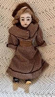 Full body shot of an 18-inch J.D. Kestner bisque doll wearing a brown wool jacket with detachable collar, matching skirt, furry headband and black shoes with some damage on toes.