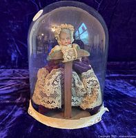 Full view of the lacemaker doll seated on wooden chair with lacemaking pillow, under large clear glass dome against purple velvet background.